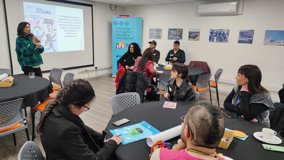 Un mujer expone ante una sala con personas sentadas en mesas redondas