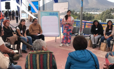 Grupo de personas sentadas en círculo escuchando a mujer que expone 
