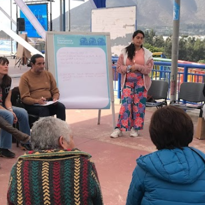 Grupo de personas sentadas en círculo escuchando a mujer que expone 